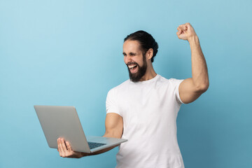 Portrait of successful man with beard wearing white T-shirt screaming with joy and holding laptop, rejoicing victory, online betting. Indoor studio shot isolated on blue background.