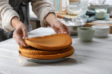 Woman stacking homemade sponge cakes at white wooden table in kitchen, closeup