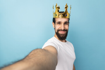 Portrait of satisfied handsome man with beard wearing white T-shirt and gold crown, looking at...