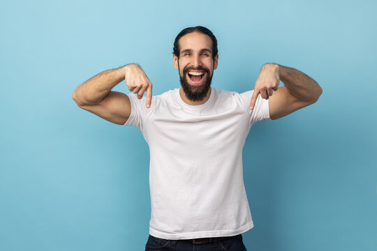 Look at ads below. Portrait of man with beard wearing white T-shirt smiling and pointing down, showing place for idea presentation, commercial text. Indoor studio shot isolated on blue background.