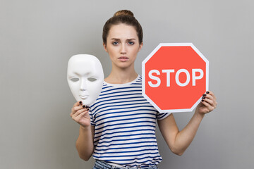 Portrait of serious woman wearing striped T-shirt holding white mask with unknown face and red...