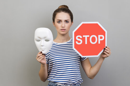 Portrait of serious woman wearing striped T-shirt holding white mask with unknown face and red traffic sign, looking at camera. Indoor studio shot isolated on gray background.