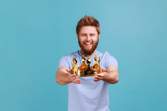 Portrait of smiling happy bearded man holding out golden crown, looking at camera with toothy smile, expressing positive emotions. Indoor studio shot isolated on blue background.