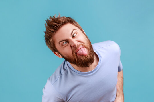 Portrait of crazy childish handsome young adult bearded man looking at camera with crossed eyes and showing tongue out, expressing positive emotions. Indoor studio shot isolated on blue background.