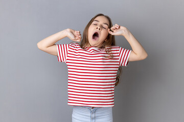 Portrait of sleepy little girl wearing striped T-shirt yawning and raising hands up, feeling...