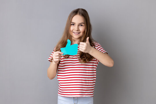 Little girl wearing striped T-shirt holding thumbs up blue icon, follower notification symbol, recommendation and good feedback in social media. Indoor studio shot isolated on gray background.