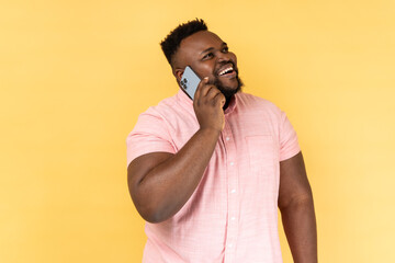 Portrait of cheerful man wearing pink shirt talking with friend on cell phone, looking away and...