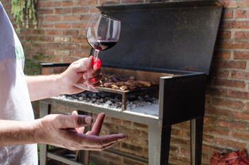 Man cooking a  traditional Argentinian barbecue, holding a glass of red wine on his hand.