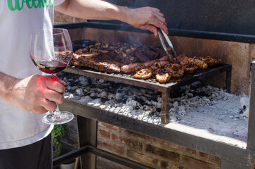 Man cooking a  traditional Argentinian barbecue, holding a glass of red wine on his hand.