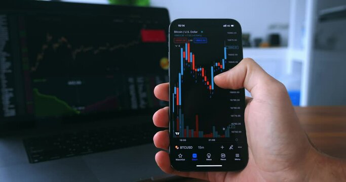 Trader working with smartphone on crypto stock exchange in front of laptop stock market financial screen at home kitchen. Trader analyzing stock market investments on smartphone app. Cryptocurrency.