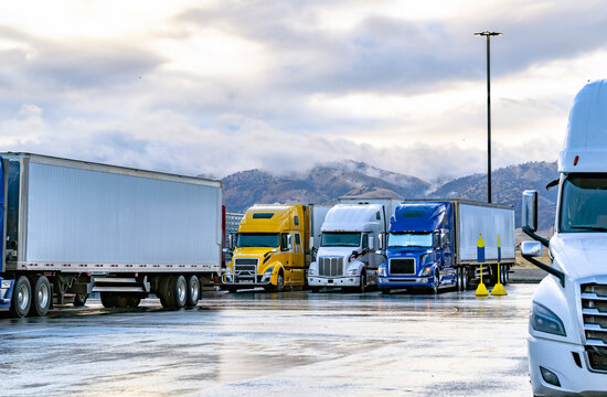 Different big rigs semi trucks with loaded semi trailers standing on the truck stop parking lot with wet surface and mountains on the background