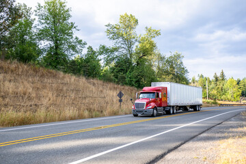 Low cab profile red big rig semi truck transporting goods in dry van semi trailer driving on the narrow mountain road