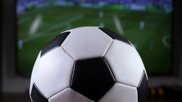 Close-up of a black and white soccer ball on the background of a TV with a football match.
