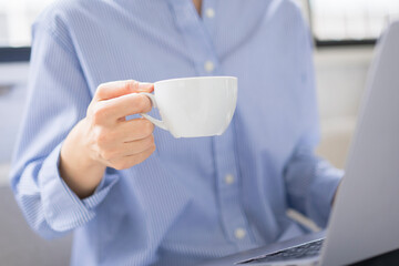 飲み物を飲みながらノートパソコンを膝に置き画面を見ているアジア人女性/白いカップを手に持っている