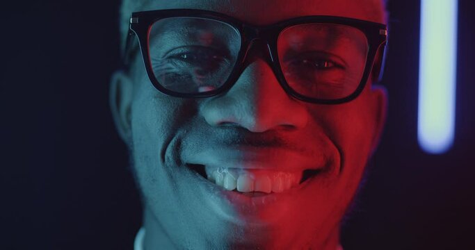 Close-up portrait of cheerful African American businessman in glasses winking and smiling on black background with neon lamps. Businesspeople and positive emotion concept.