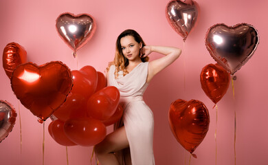 Beautiful girl with curled hair and makeup in a long pink dress on a background of heart balloons. March 8, the day of all women, a holiday.