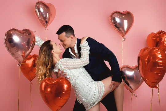 A couple in evening dresses are kissing while dancing against a background of red and pink balloons in the shape of a heart on a pink background. Vespers on Valentine's Day or March 8, Women's Day.