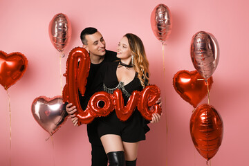 Couple in black suits holding love inscription, red balloons, pink background. A woman in a corset...