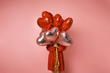 The girl is holding red and pink balloons in front of her on a pink background in a red dress. Young woman on birthday holiday party. Card post Valentine's Day, February 14.