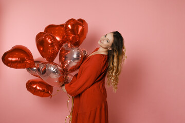 Beautiful girl in a red dress on a pink background, red and pink heart balloons. Joyful model having fun, celebrating, playing dances. Valentine's Day, February 14