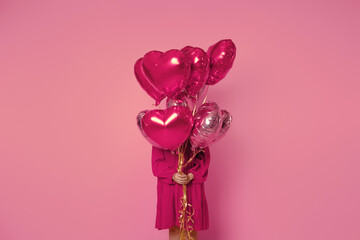 Fototapeta premium A girl holds pink balloons in front of her on a pink background in a pink dress. Young woman on birthday holiday party. Valentine's Day, February 14.