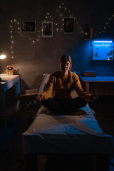 Young woman meditating on bed in lotus position sitting in dark room at night