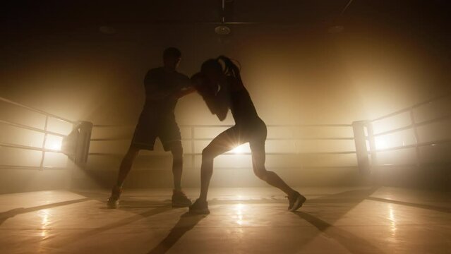 Silhouettes of man and woman having intensive workout indoors. Close-up view of female kickboxer training with her coach at boxing club. High quality 4k footage in golden orange yellow foggy light