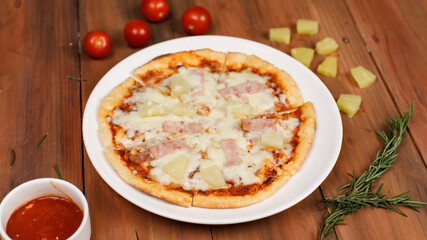 Closeup top view studio shot of round hot oven baked tasty delicious Italian homemade ham pineapple original Hawaiian soft bread crust pizza on wooden plate pan with tomatoes and ketchup on table