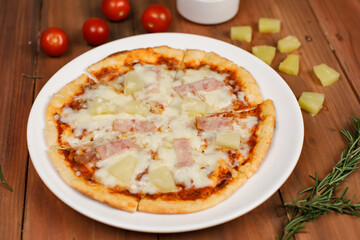 Closeup top view studio shot of round hot oven baked tasty delicious Italian homemade ham pineapple original Hawaiian soft bread crust pizza on wooden plate pan with tomatoes and ketchup on table