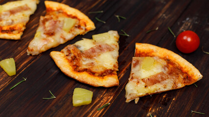 Closeup top view studio shot four pieces of hot oven baked tasty delicious Italian homemade ham and pineapple traditional Hawaiian soft bread crust pizza decorated on wooden table with tomatoes herbs