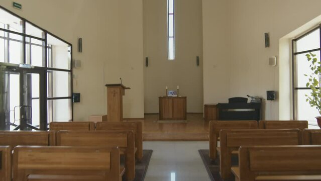 No people shot of small Catholic church interior, camera moving along nave between rows of wooden pews to bema with altar and cross shaped window