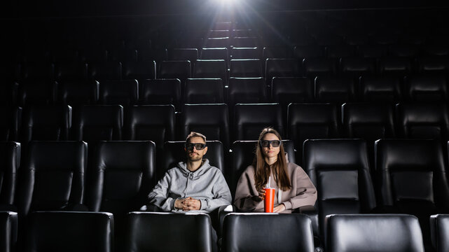 A couple in love is watching a movie with 3D glasses and drinking from a red cup. Cinema for two - Powered by Adobe