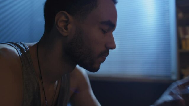 Handheld side view close-up of young Black man suffering depression sitting in bedroom drinking whiskey late at night