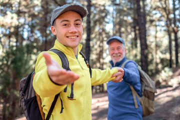 Naklejka premium Handsome young boy walks happily in the forest holding his grandfather's hand. Smiling multigenerational family enjoying freedom mountain and nature together. The new generation helps the old one