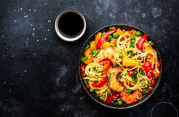 Stir fry noodles with shrimps, red and yellow paprika, green pea, chives and sesame seeds bowl. Asian cuisine dish. Black stone kitchen table background, top view