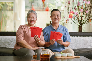Happy senior couple sitting on couch at home and showing red envelopes prepared for Tet