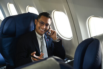 Confident businessman in black suit sitting comfortable seat in airplane cabin and talking on mobile phone