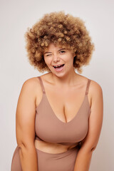 Vertical shot of plus size body positive adult woman with curly hair in lingerie winks to the camera playfully and smiles widely, poses at the studio against white background. Body positive and self