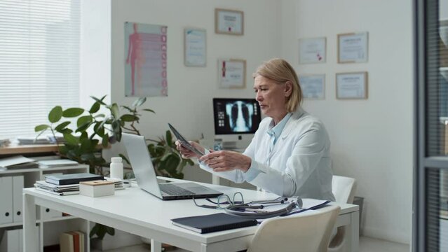 Mature radiologist sitting by workplace in front of laptop, pressing button on keyboard, taking lung x-ray of online patient and looking at image during remote consultation