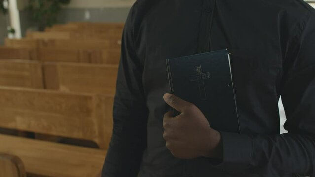 Medium section tracking shot of unrecognizable Catholic priest holding the Bible walking along church nave