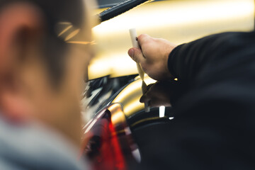 Rear view of man removing dents from body of car using bright lamp. Professional car repair and detailing in a garage. Blurred foreground. Horizontal close-up indoor shot. High quality photo