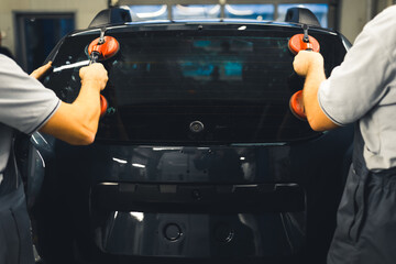 Rear view of two men installing rear window pane into back of car using professional tools. Garage...