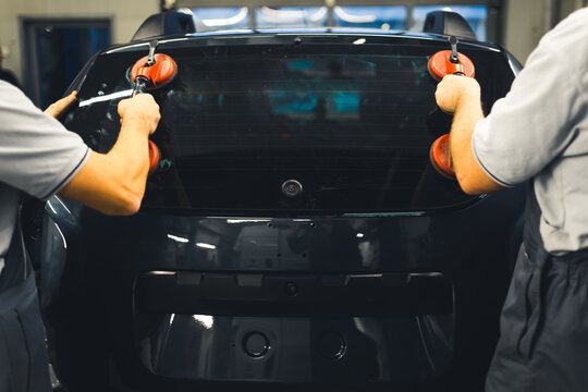 Rear View Of Two Men Installing Rear Window Pane Into Back Of Car Using Professional Tools. Garage Work. Car Maintenance. Horizontal Indoor Shot. High Quality Photo