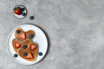 Heart shaped pancakes for romantic breakfast. Valentine's Day concept. Table scene with a white plate, strawberry and blueberry. Grey background. Top view. Love and hearts theme. Copy space