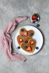 Heart shaped pancakes for romantic breakfast. Valentine's Day concept. White plate, pink cloth napkin, metal fork and knife, fresh berries. Grey background. Top view. Love and hearts theme