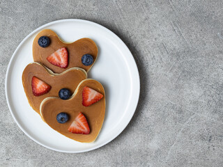 Three heart shaped pancakes for romantic breakfast. Valentine's Day concept. Table scene with a white plate, strawberry and blueberry. Grey background. Top view. Love and hearts theme. Copy space