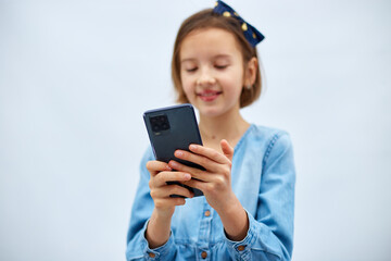 Smiling little girl in casual denim dress hold smartphone, using mobile cell phone internet applications, typing message isolated on white background, studio. Child and modern tech usage concept