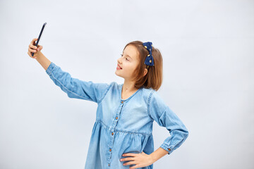 Smiling little girl in casual denim dress hold smartphone do selfie shot on mobile cell phone isolated on white background, studio.  lifestyle concept.