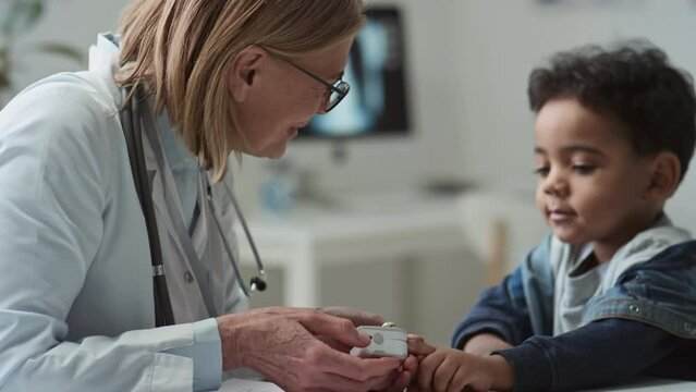 Experienced female pediatrician putting pulse oximeter on fingertip of cute little patient to check level of oxygen saturation in his blood while young man signing medical document
