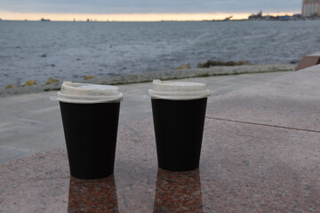 Two paper cups for coffee stand at a distance from each other on a marble border against the blue sea. A takeaway cup with a lid with a ready drink. Holiday concept for two, recycling of used material